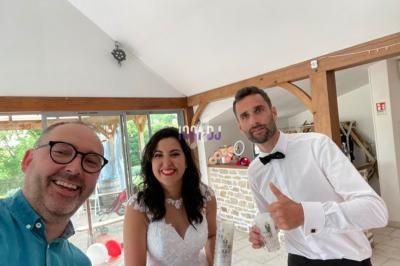 Un couple en tenue de mariage pose avec un homme souriant dans une salle lumineuse au décor moderne et rustique.