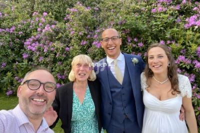 Quatre personnes souriantes posent devant des buissons fleuris, dont un couple en tenue de mariage.