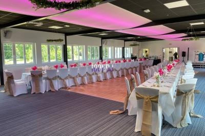 Salle de réception décorée avec des tables alignées, nappes blanches, serviettes roses et chaises ornées de rubans beiges.