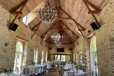 Salle de réception en pierre avec plafond en bois, lustres suspendus et tables dressées pour un événement.