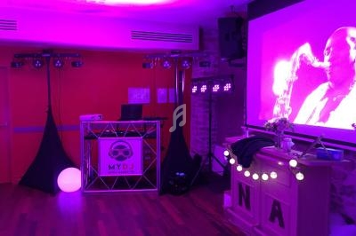Salle de réception éclairée en lumière violette avec un stand de DJ, un écran projetant un saxophoniste et des décorations…