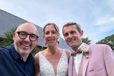 Un homme souriant prend un selfie avec un couple habillé en tenue de mariage devant un bâtiment avec un toit en ardoise.
