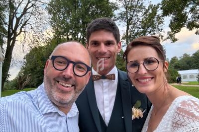 Un homme prend un selfie avec un couple souriant en tenue de mariage dans un parc verdoyant.