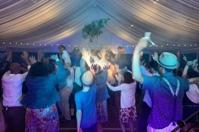 Des personnes dansent sous une tente éclairée de guirlandes lumineuses, bras levés, dans une ambiance festive.