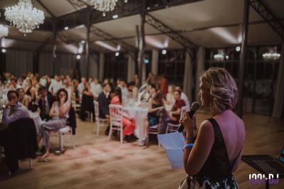 Une femme fait un discours au micro devant un public assis dans une salle éclairée par des lustres.