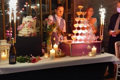 Un homme remplit une pyramide de coupes avec du champagne lors d'une réception festive, entouré de décorations lumineuses.