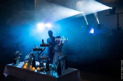 Un couple se tient devant une table décorée, éclairée par des lumières de scène dans une ambiance enfumée.