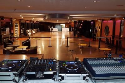 Salle de réception vide avec une scène équipée de matériel de mixage audio et des tables dispersées.