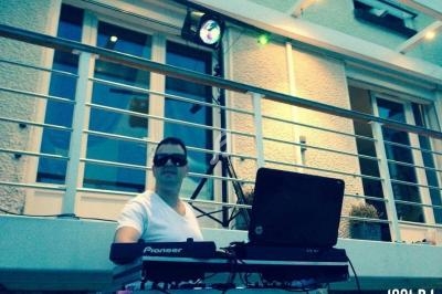 Un DJ mixe en extérieur devant une maison, équipé d'un ordinateur portable et de matériel audio.