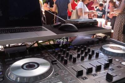 Table de mixage avec ordinateur en premier plan, foule de personnes discutant en extérieur sous un ciel ensoleillé en…