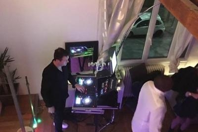 Un DJ mixe de la musique sur une console dans une pièce éclairée, avec des personnes dansant à proximité.