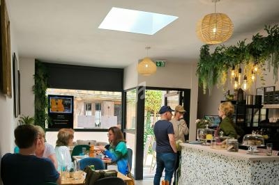 Intérieur d'un café lumineux avec des clients discutant et une serveuse derrière un comptoir décoré de plantes.