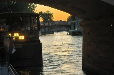 Vue d'un bateau amarré sous un pont au coucher du soleil, avec une rivière et des arbres en arrière-plan.