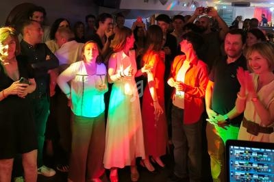 Groupe de personnes debout dans une ambiance festive, éclairées par des lumières colorées, avec un DJ en arrière-plan.