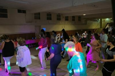 Des personnes dansent en groupe dans une salle éclairée par des lumières colorées, avec des tables et spectateurs en arrière…