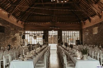 Grande salle en pierre avec tables longues dressées pour un repas, éclairée par des guirlandes lumineuses suspendues.