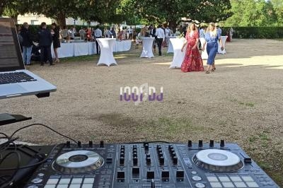 Console de DJ en premier plan avec des invités debout et discutant dans un jardin lors d'un événement en extérieur.