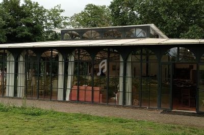 Serre en verre et métal avec toit incliné, entourée de verdure, utilisée comme espace intérieur aménagé.