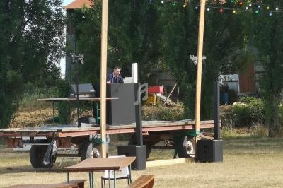 Scène extérieure en bois avec un DJ en arrière-plan, entourée de tables et bancs sur une pelouse.