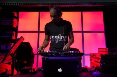 Un DJ mixe sur une console dans une ambiance lumineuse avec un fond rose et rouge.