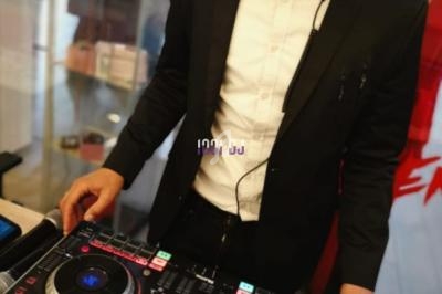 Un DJ en costume noir et nœud papillon rouge mixe sur une table de mixage dans un environnement intérieur.