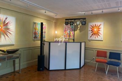 Salle d'exposition avec des peintures colorées accrochées aux murs, un espace DJ et des chaises disposées.