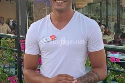 Un DJ souriant mixe de la musique en extérieur, devant une table de mixage, entouré de fleurs et de lumières.