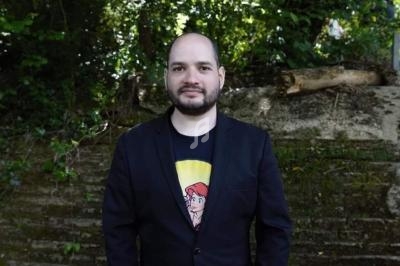 Homme debout devant un mur en pierre, vêtu d'une veste noire et d'un t-shirt imprimé, entouré de végétation.