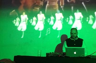 Un DJ mixe sur son ordinateur devant une projection murale de danseurs en tenue blanche.