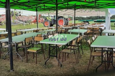 Tables et chaises disposées sous des tentes vertes dans un champ, avec des tracteurs en arrière-plan.