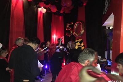 Groupe de personnes célébrant un anniversaire dans une salle décorée en rouge et noir avec des ballons marqués ’50’.