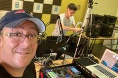 Un homme souriant devant une console de mixage dans un studio radio, une autre personne en arrière-plan.