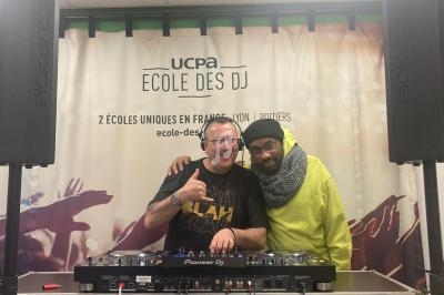Deux personnes souriantes posent devant une table de mixage, avec une bannière ’UCPA École des DJ’ en arrière-plan.