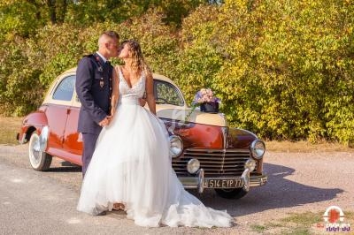 Un couple en tenue de mariage pose devant une voiture ancienne rouge et beige, garée près d'arbres.