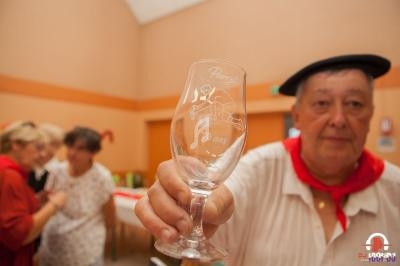 Un homme en béret montre un verre gravé ’Pierrot 70 ans’ dans une salle avec des personnes en arrière-plan.