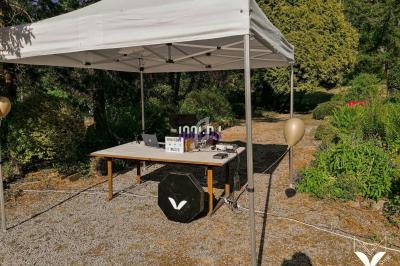 Stand extérieur sous une tente blanche, équipé d'une table avec ordinateurs et matériel, situé dans un jardin.