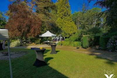 Jardin avec pelouse, tables hautes noires et tentes blanches, entouré d'arbres et de végétation.