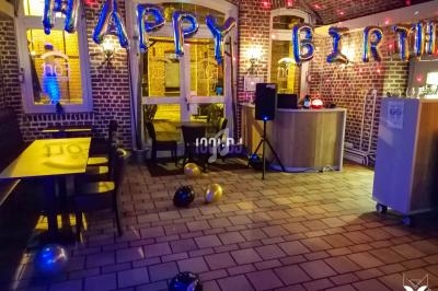 Salle décorée pour un anniversaire avec des ballons ’Happy Birthday’, des tables, des chaises et un bar en arrière-plan.