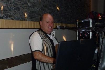 Homme souriant debout derrière un ordinateur portable, tenant un micro dans un environnement intérieur.