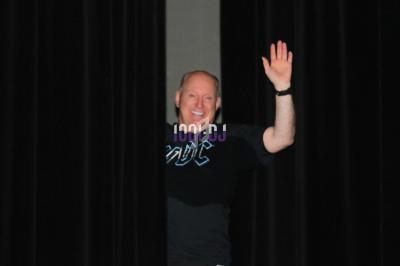 Un homme souriant, vêtu d'un t-shirt noir, salue en apparaissant entre deux rideaux noirs.