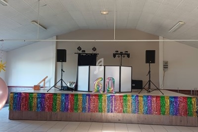 Scène décorée avec des guirlandes colorées, ballons et chiffres ’60’, équipée de matériel audio et éclairage.