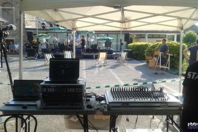 Matériel de sonorisation et de mixage installé sous un barnum, avec une scène en arrière-plan dans une cour extérieure.