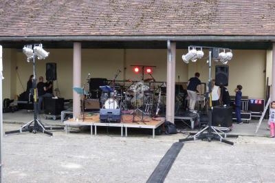 Scène extérieure avec instruments de musique installés, éclairages suspendus et techniciens en préparation.