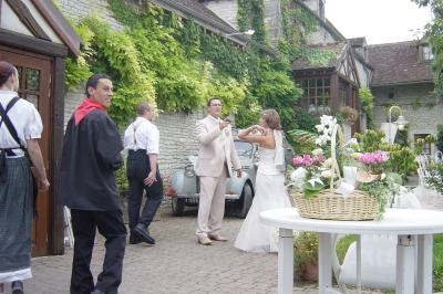 Un couple de mariés échange un toast devant une bâtisse en pierre entourée de végétation, avec des invités en arrière-plan.