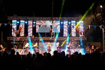Groupe de musiciens et danseuses sur une scène illuminée lors d'un concert en plein air, devant un public.