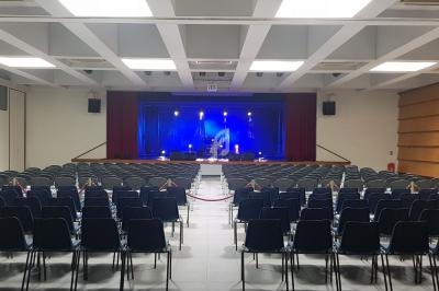 Salle de spectacle vide avec des rangées de chaises noires face à une scène éclairée par des projecteurs bleus.