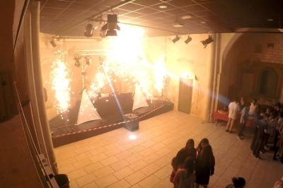 Effets pyrotechniques illuminant une petite scène dans une salle en pierre, avec des spectateurs rassemblés à droite.