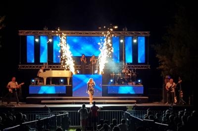 Scène de concert nocturne avec une chanteuse au centre, des musiciens et des effets pyrotechniques en arrière-plan.