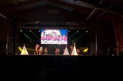 Spectacle sur scène avec danseurs en costumes traditionnels, décor de tipis et éclairage coloré dans une salle sombre.