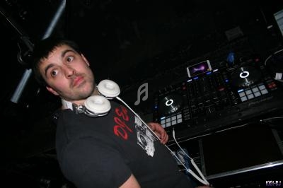 Un DJ portant un casque autour du cou se tient près d'une table de mixage dans un environnement sombre.